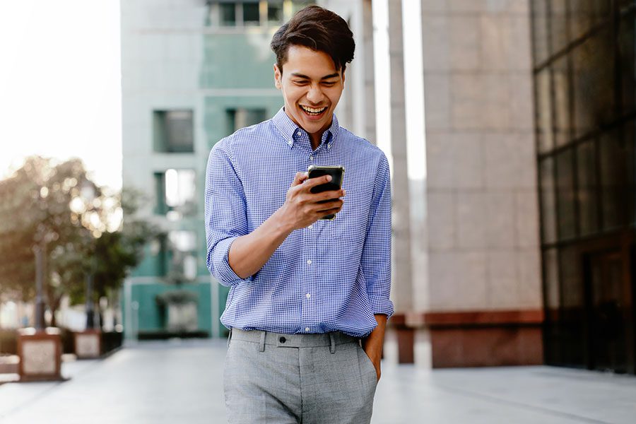 Client Center - Smiling Young Businessman Walking Outside a Modern Commercial Building While Using a Cellphone