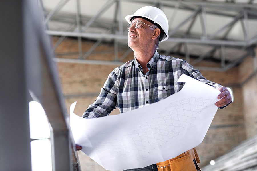 Specialized Business Insurance - View of Smiling Senior Contractor Looking Up at a New Building Under Construction While Holding Blueprints in His Hands